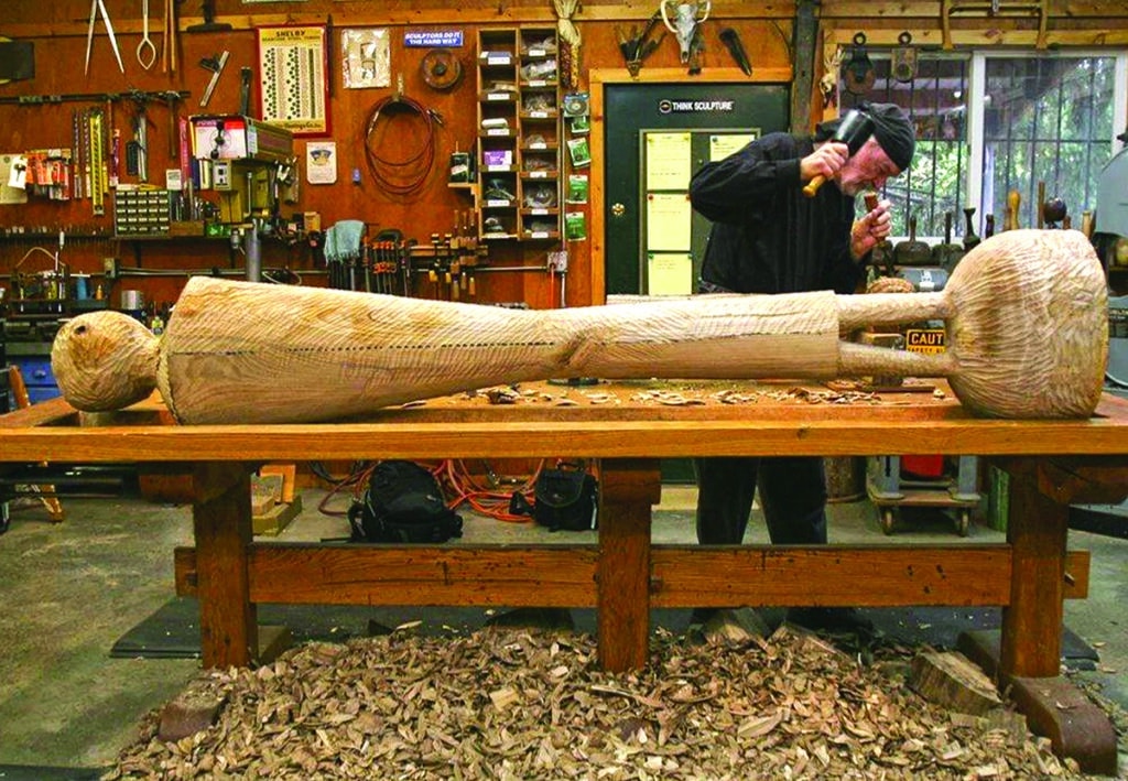 David Hostetler using a mallet and gouge to hand-carve a large reclining female figure from a log in his studio