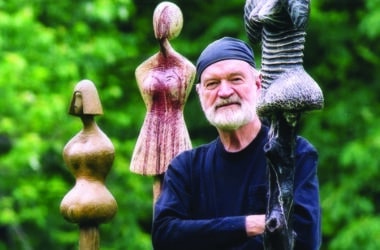 David Hostetler standing among his wood-carved female sculptures in an outdoor garden setting