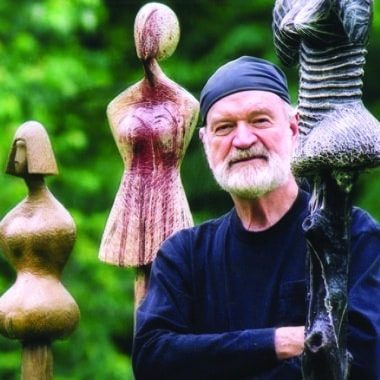 David Hostetler standing among his wood-carved female sculptures in an outdoor garden setting