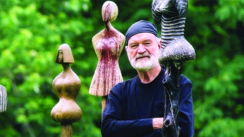 David Hostetler standing among his wood-carved female sculptures in an outdoor garden setting