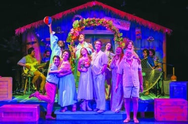 The full cast of Escape to Margaritaville at Theatre Workshop of Nantucket celebrating on a colorful tropical beach stage set