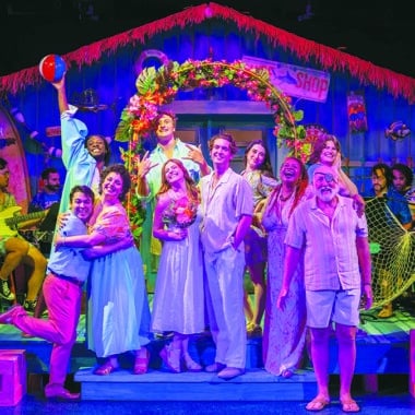 The full cast of Escape to Margaritaville at Theatre Workshop of Nantucket celebrating on a colorful tropical beach stage set