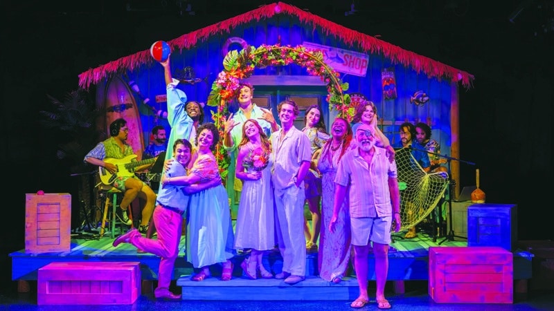 The full cast of Escape to Margaritaville at Theatre Workshop of Nantucket celebrating on a colorful tropical beach stage set