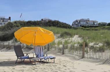 Beach chairs and a yellow umbrella on a sunny Nantucket beach with sand dunes, beach grass, and classic shingled homes in the background