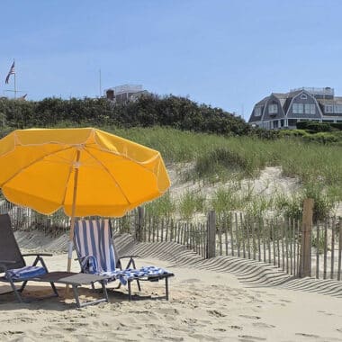 Beach chairs and a yellow umbrella on a sunny Nantucket beach with sand dunes, beach grass, and classic shingled homes in the background