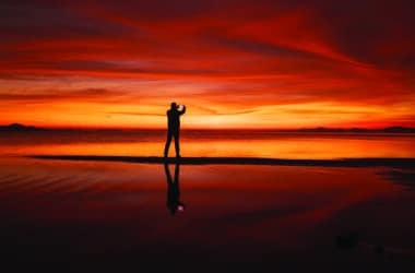 Alt text: A silhouetted figure photographs a vivid red and orange sunset reflected on wet sand, capturing the tension between experiencing a place and posting it on social media.