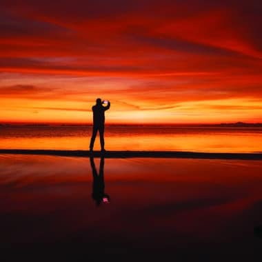 Alt text: A silhouetted figure photographs a vivid red and orange sunset reflected on wet sand, capturing the tension between experiencing a place and posting it on social media.