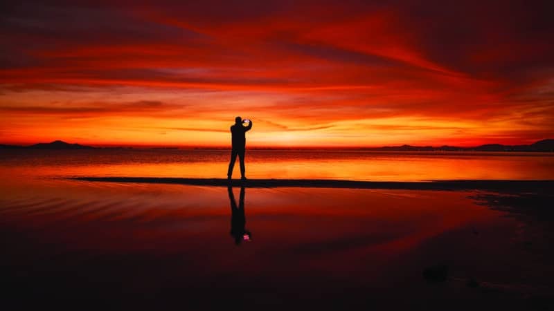 Alt text: A silhouetted figure photographs a vivid red and orange sunset reflected on wet sand, capturing the tension between experiencing a place and posting it on social media.