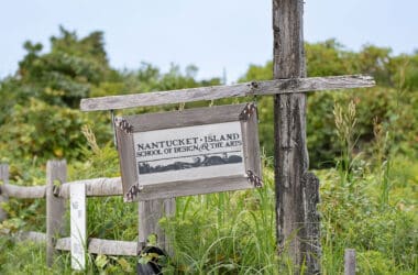A weathered wooden sign for the Nantucket Island School of Design and the Arts hangs on a post surrounded by lush green meadow vegetation at Seaview Farm.