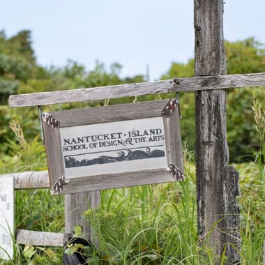 A weathered wooden sign for the Nantucket Island School of Design and the Arts hangs on a post surrounded by lush green meadow vegetation at Seaview Farm.