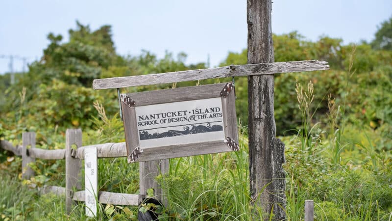 A weathered wooden sign for the Nantucket Island School of Design and the Arts hangs on a post surrounded by lush green meadow vegetation at Seaview Farm.