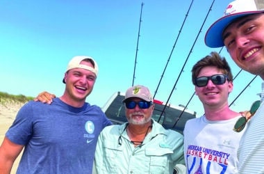 Nantucket fishing guide Steve Tuna Tornovish poses with three clients on a sunny beach with surf fishing rods loaded on a truck behind them.