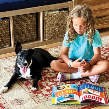 A young girl reads a picture book with her dog beside her, reflecting the love of reading Nantucket Children's Book Day hopes to inspire.