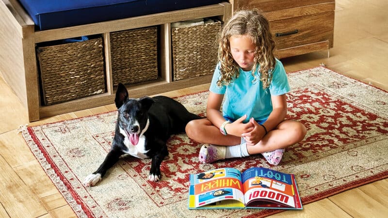 A young girl reads a picture book with her dog beside her, reflecting the love of reading Nantucket Children's Book Day hopes to inspire.