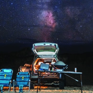 The Milky Way blazes over a Nantucket beach camp setup, the kind of dark sky the essay describes discovering at night.