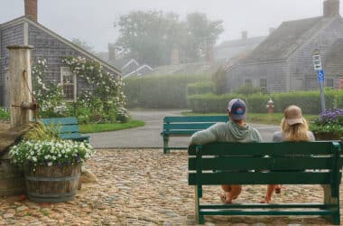 Two people sitting on a green bench overlooking a cobblestone street and historic Nantucket cottages on a foggy day.