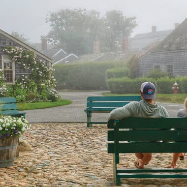 Two people sitting on a green bench overlooking a cobblestone street and historic Nantucket cottages on a foggy day.