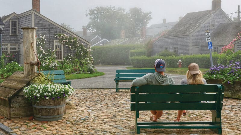 Two people sitting on a green bench overlooking a cobblestone street and historic Nantucket cottages on a foggy day.